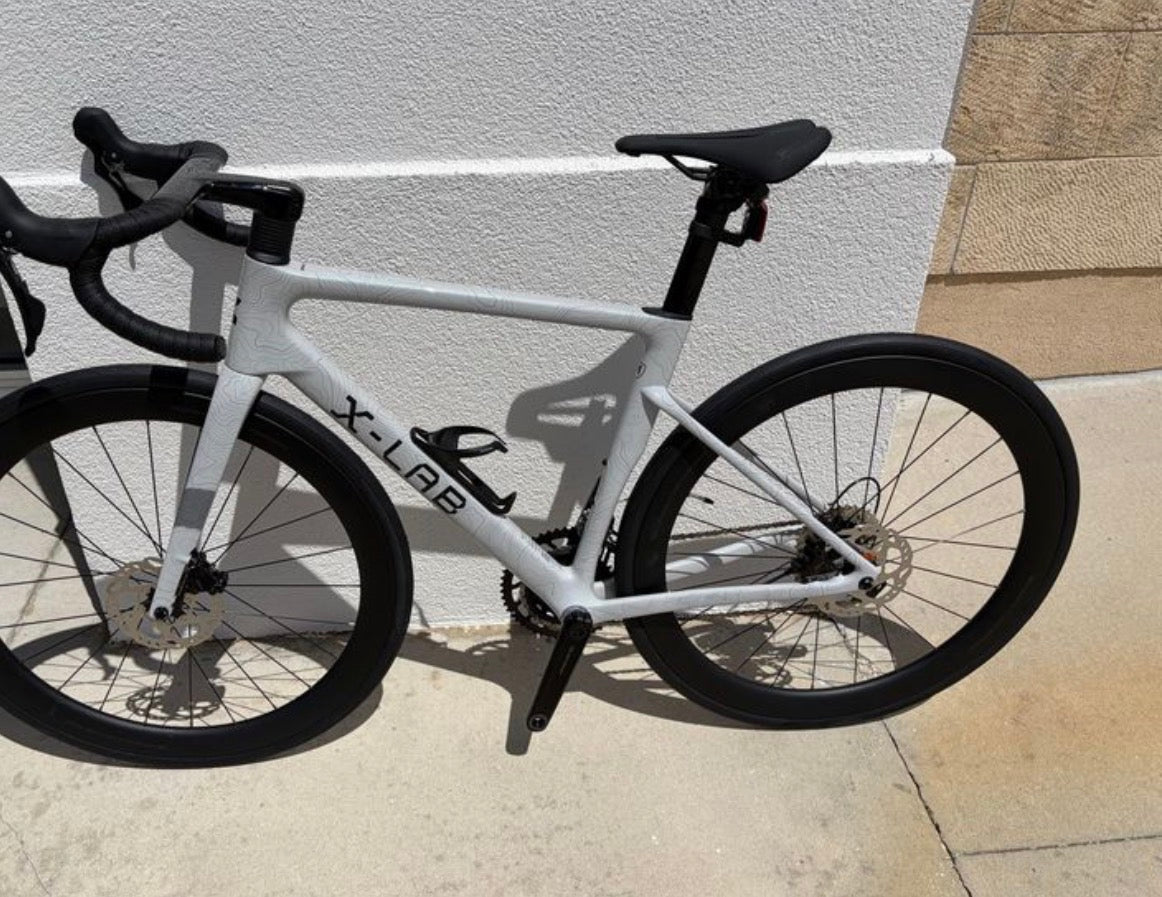 A white X-LAB road bicycle with black components, including the saddle, handlebars, wheels, and drivetrain, photographed outdoors against a light-colored stucco wall.