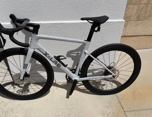 A white X-LAB road bicycle with black components, including the saddle, handlebars, wheels, and drivetrain, photographed outdoors against a light-colored stucco wall.